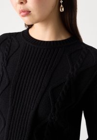 Femme portant un pull en maille noir texturé et une seule boucle d'oreille métallique pendante, montrée des épaules au menton, face à gauche.