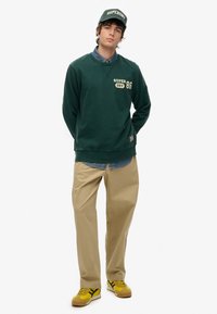 Zielony sweatshirt z logo "SUPERDRY 86", khaki spodnie i żółte sneakersy. Pod spodem koszula dżinsowa oraz pasująca czapka. Luźny krój.