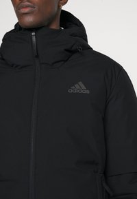 Svart Adidas-jacka med hög krage och huva, med dragkedjestängning, texturerat material och en diskret logo på bröstet.