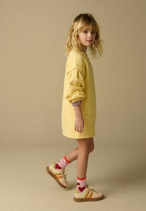 Jong meisje met blond haar, gekleed in een gele oversized trui, kleurrijke sokken met hartjespatroon en beige sneakers, lopend zijwaarts.