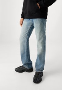 Jeans de ganga azul claro com corte reto, apresentando áreas desbotadas, design de cinco bolsos e textura de tecido desgastado, combinados com ténis pretos.