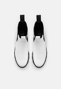 Botas de tobillo de cuero blanco con acentos laterales de elasticidad negra y suela negra texturizada. Presentan una tirilla en el talón para facilitar su uso.