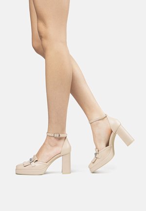 Scarpe con tacco alto beige con platform, cinturino alla caviglia e dettaglio a fiocco. Materiale in pelle liscia con punta quadrata e design a tacco largo.