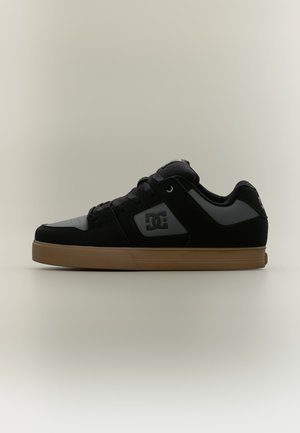 Μαύρο και γκρι sneaker χαμηλού προφίλ με ταμπά σόλα, σχέδιο κορδονιών και λογότυπο DC στο πλάι, εμφανίζεται σε ουδέτερο φόντο.