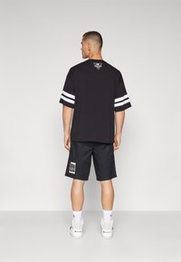 Camiseta negra de gran tamaño con rayas blancas en las mangas; combinada con pantalones cortos negros que tienen una etiqueta en el lado, llevada con zapatillas blancas.