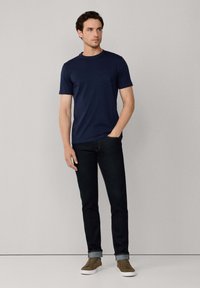 Camiseta de algodón color azul marino con mangas cortas y cuello redondo, que presenta un pequeño bolsillo delantero. Combinada con jeans de mezclilla oscuros y zapatillas marrones.