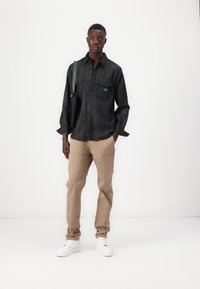Black denim shirt with button front, two chest pockets, tan chinos, and white sneakers, standing against a plain white background.