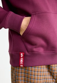 Mano descansando dentro de un bolsillo de sudadera color burdeos con una etiqueta roja que dice "RETIRAR ANTES" sobre pantalones de cuadros marrones y azules.