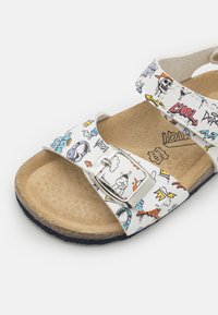 Hvit sandal med fargerik grafikk, spennedetalj, korkfotseng og teksturert svart gummisåle.