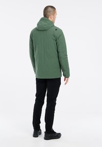 Chaqueta aislante verde con capucha, que presenta una textura suave, puños elásticos y un diseño sencillo. Se lleva con pantalones y zapatos negros.