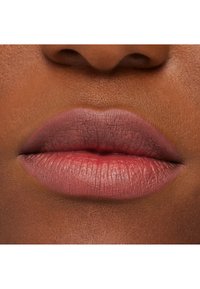 Primer plano de labios con un color rosa mate y apagado. La textura es suave, con líneas sutiles visibles, en contraste con un tono de piel marrón más profundo.