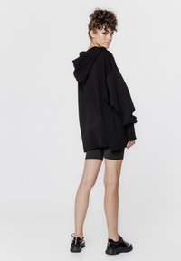 Czarna, oversize bluza z kapturem, z opadającymi ramionami i długimi rękawami, w połączeniu z ciemnymi szortami rowerowymi i czarnymi trampkami.