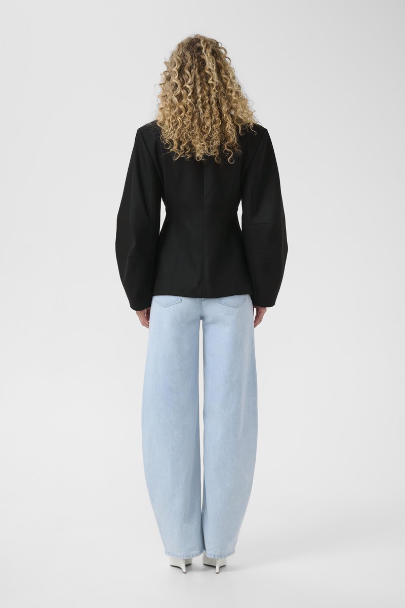 Personne aux cheveux bouclés portant une veste noire ajustée à larges manches, un jean bleu clair à jambes larges et des chaussures à talons blanches, debout face à l'arrière.