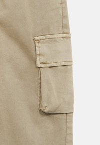 Bež hlače cargo z izrazitim stranskim žepom, izdelane iz trpežne mešanice bombaža z teksturirano diagonalno rebrasto tkanino.