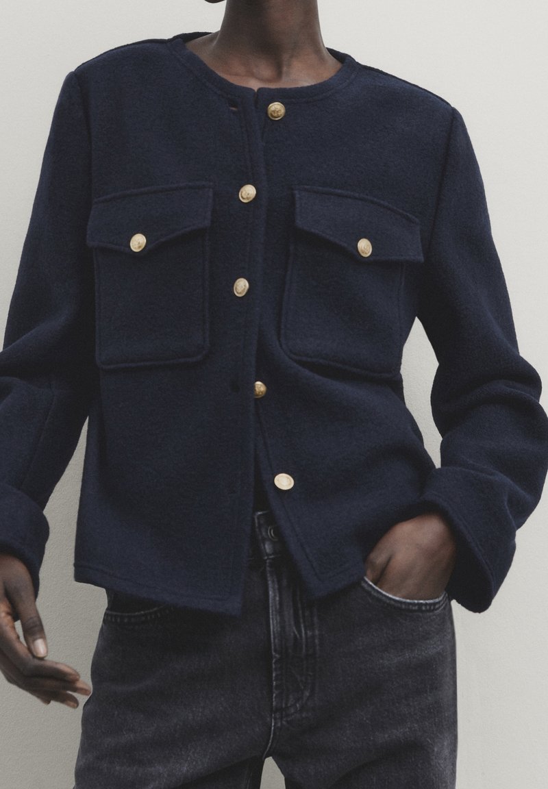 Personne à la peau foncée portant une veste bleu marine à boutons avec quatre boutons dorés et deux poches poitrine, associée à un jean noir, debout contre un mur blanc.