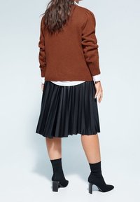 Femme portant un pull en tricot marron, une chemise blanche, une jupe midi plissée noire et des bottines chaussettes à talons noires, vue de dos.