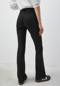 Pantaloni negri evazați cu un brâu elastic, textură netedă și design ajustat, asortați cu adidași întunecați care au accente albe.