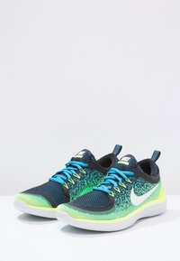 Černé a zelené sportovní boty s textilním vrškem z prodyšné látky, modrými tkaničkami, bílým logem ve tvaru „swoosh“ a texturovanou podešví. Obsahují poutko vzadu.