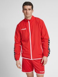 Rød atletisk jakke med fuld lynlås, elastiske manchetter og sort chevron mønster på ærmerne, parret med røde shorts med hvid kant.