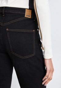 Los jeans negros presentan un parche de cuero marrón con la marca "BOSS", costuras amarillas en contraste y un diseño ajustado con bolsillos traseros.