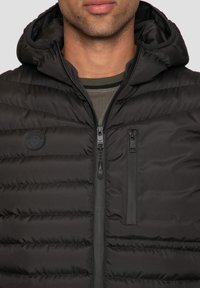 Veste matelassée noire avec capuche, présentant une texture matelassée horizontale, une fermeture éclair à l'avant et une poche poitrine. Fabriquée en nylon.