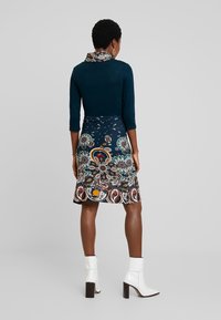 Klänning med en mörk teal topp med krage och en mönstrad kjol i blått, vitt och flerfärgade blommönster. Vita ankelskor.