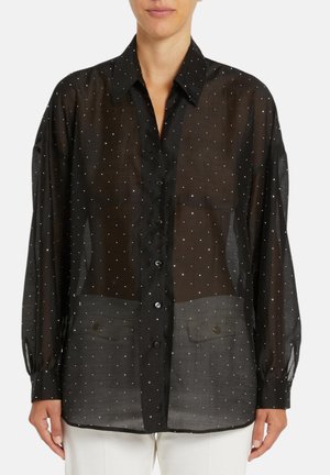 Blusa nera trasparente a maniche lunghe con pois bianchi, chiusura con bottoni sul davanti, colletto classico e due tasche frontali con pattina, indossata sopra pantaloni bianchi.