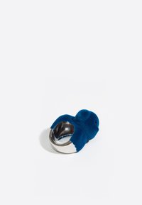 Sudraba gredzens ar strukturētu zilu samta vaļa figūru, parādīts uz baltas fona.