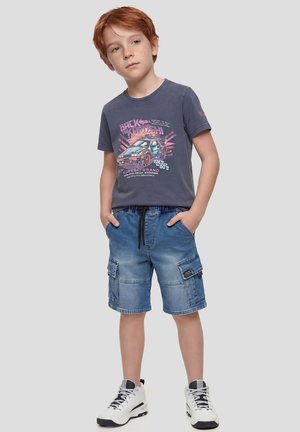 Niño pelirrojo de pie con las manos en los bolsillos, vistiendo una camiseta gráfica oscura, pantalones cortos cargo azules y zapatillas blancas sobre un fondo gris.