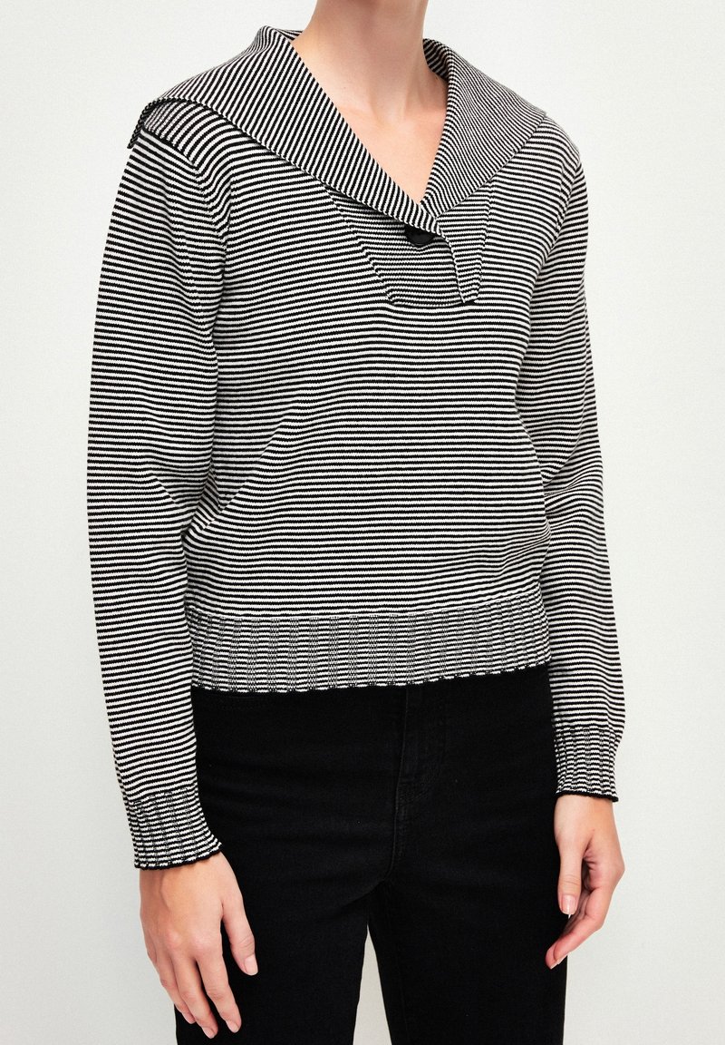 Maglione a righe bianche e nere con un ampio colletto da marinaio, maniche lunghe e polsini a costine. Presenta un dettaglio con bottone sul colletto.