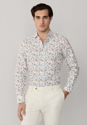 Homme portant une chemise blanche à manches longues boutonnée, ornée d'imprimés colorés d'animaux et de plantes, associée à un pantalon beige clair.