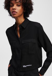 Camicia nera con maniche lunghe e bottoni, realizzata in tessuto liscio. Presenta due tasche sul petto e un logo sull'orlo. Design corto.
