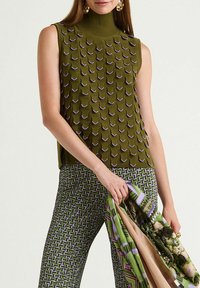 Haut vert sans manches avec un motif texturé et des accents violets. Associé à un pantalon à motifs verts et violets, tenant une écharpe multicolore.