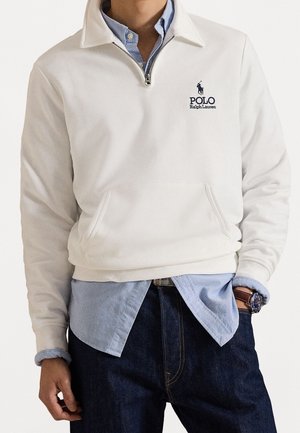 Person iført en hvid Polo Ralph Lauren trøje med kvartlynlås over en blå skjorte med knapper og mørke jeans, med et ur synligt.
