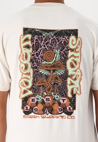 Camiseta de algodón beige con un diseño gráfico colorido que presenta un caracol, champiñones y hexágonos. El texto dice "Vulcan Store EST 1991."