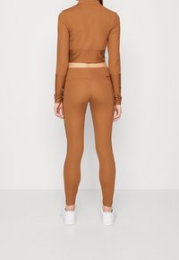 Hnědý krátký top s dlouhým rukávem a odpovídající legíny z hladkého, elastického materiálu. Bílé tenisky dokončují celkový vzhled.