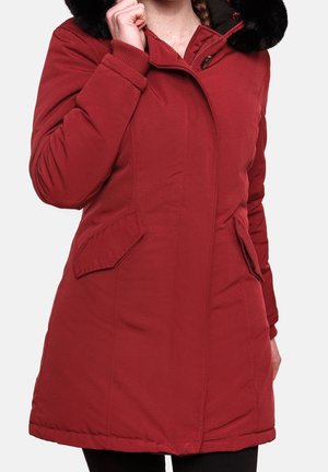 Czerwona ocieplana zimowa kurtka z kieszeniami na patki i czarnym kapturem wyściełanym futrem, noszona przez osobę poprawiającą kołnierz.