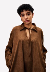 Frau mit kurzen lockigen Haaren, die ein lockeres braunes Wildlederoberteil mit Kragen und vorderem Knoten trägt, steht mit Händen in den Taschen vor einem weißen Hintergrund.