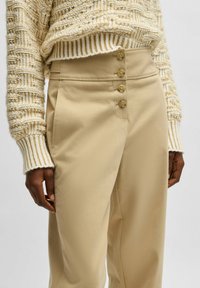 Beige högmidjade byxor med en slät textur, med fyra dekorativa knappar i midjan och sidofickor. Tröjan har ett stickat mönster.