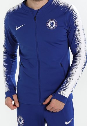 Niebieski dres Chelsea FC z białymi wzorami przypominającymi pociągnięcia pędzla na rękawach, zapinana na zamek kurtka oraz dopasowane spodnie z logo klubu i Nike.
