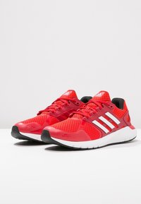 Zapatillas deportivas rojas con parte superior de malla, logo de tres rayas blancas, cuello acolchado y suela ligera. Presenta detalles texturizados y cordones.