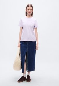 Ανοιχτό μωβ βαμβακερό t-shirt με κοντά μανίκια και μικρό λογότυπο· συνδυασμένο με σκουρόχρωμη τζιν φούστα με μπροστινή σχισμή. Μπορντό παπούτσια και κρεμ τσάντα.