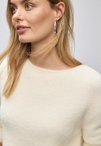 Cremfarbener, flauschiger Pullover mit weitem Ausschnitt und weicher Textur. Silberne Creolen betonen den Look. Haare locker gestylt.