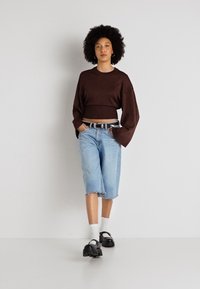 Sweater castanho curto com mangas largas, combinado com calções de ganga de comprimento até ao joelho em azul claro. Sapatos grossos pretos e meias brancas completam o look.