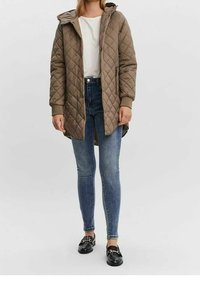 Chaqueta acolchada con capucha de color marrón claro, con puños y dobladillo acanalados, combinada con jeans ajustados azules y zapatos negros con detalles dorados.