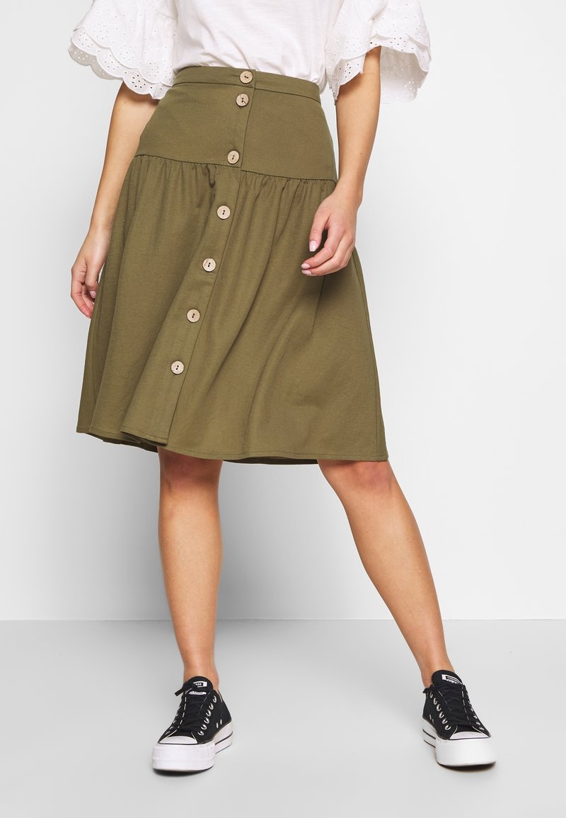 Jupe longueur genoux vert olive avec une taille cintrée, un bas évasé et six boutons en bois sur le devant. Associée à des baskets noires.
