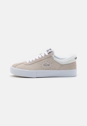 Baskets en daim beige avec semelle en caoutchouc blanche, dotées de lacets blancs, de détails perforés et d'un petit logo Lacoste sur le côté.