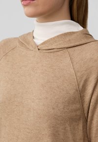 Bruine hoodie met een zachte textuur, voorzien van een diepe halslijn en een subtiel, gevlekt patroon, gedragen over een witte coltrui.