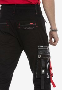 Schwarze Cargo-Hosen mit roten Nähten, mehreren Taschen und Lederakzenten mit Nieten. Logoprint auf den Gesäßtaschen und seitlichen Riemen.