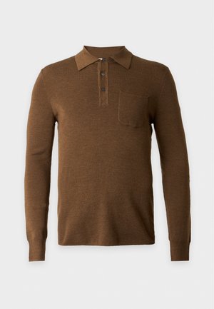 Un homme est debout face à l'objectif, vêtu d'un polo marron à manches longues, d'un pantalon beige et de baskets blanches sur un fond clair uni.